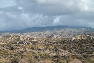 The arid landscape of Tabernas Desert in Almeria, Spain, with rugged hills, sparse vegetation, and rocky outcrops under a cloudy sky. It’s a popular filming location
