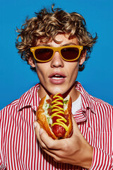 Stylish young man wearing sunglasses in colorful outfit holding a hot dog with mustard. Funny and trendy portrait on blue background