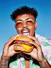 Young man wearing sunglasses eating a big cheeseburger under blue sky. Stylish, trendy and fun lifestyle portrait for food marketing