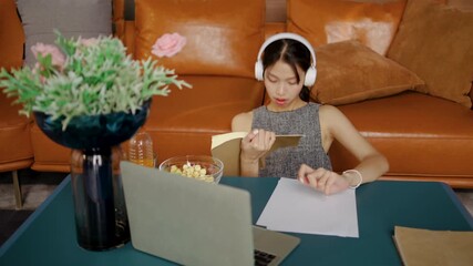 Asian female student enjoys listening to calming music through headphones while engrossed in reading a textbook and and use a laptop computer to find information in her inviting study environment