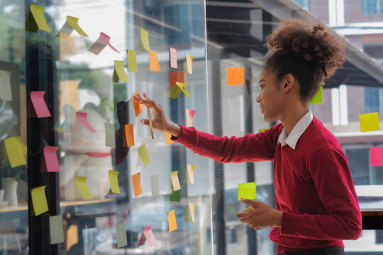 An African American businesswoman is using sticky notes on a glass wall in her office to brainstorm and develop a new strategy. - Powered by Adobe