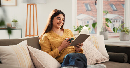 Obraz premium Woman, reading book and coffee on sofa, happy and relax with literature in living room at apartment. Person, smile and novel on couch in lounge for beverage, poetry and fiction story at house