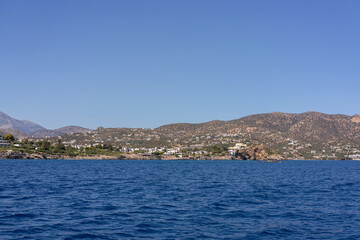 Fototapeta premium Panoramic view of Agios Nikolaos town on the coast of Crete, Greece, captured from the sea on a sunny day. The image features blue Mediterranean water, distant mountains, and the picturesque urban