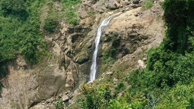 A stunning view of a waterfall located at Sawangan Beach in Kebumen, Central Java. The waterfall flows gracefully down a rocky cliff surrounded by lush green vegetation, creating a beautiful contrast.