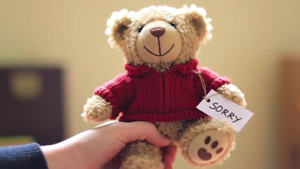 Hand holding teddy bear with 'sorry' tag, soft background, for apology or empathy themes