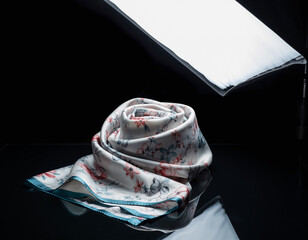 Elegant silk scarf rests on a reflective black surface under studio lighting, showcasing its delicate floral pattern and soft texture in a sophisticated manner.