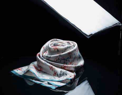 Elegant silk scarf rests on a reflective black surface under studio lighting, showcasing its delicate floral pattern and soft texture in a sophisticated manner. - Powered by Adobe