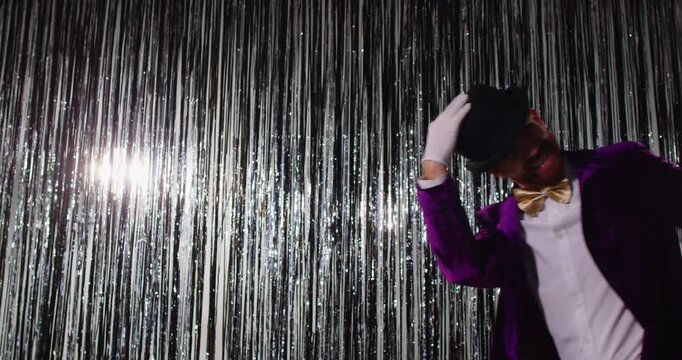 Mysterious man pretending being magician, wearing black hat, purple costume and white gloves, appearing over shiny silver background, participating in performance during party on holiday