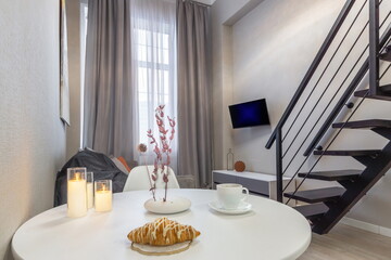 A comfortable living area features a small table with a croissant and coffee. Soft lighting from candles creates a cozy atmosphere near a modern staircase and stylish decor