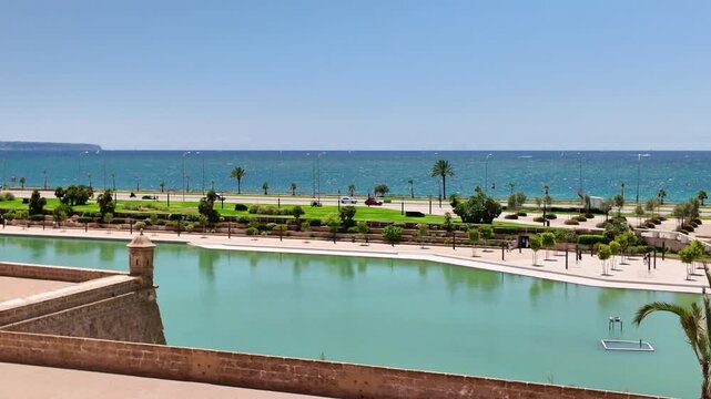 Parc de la Mar with its large saltwater lake in front of the ancient city walls, with the Mediterranean Sea and a coastal road with traffic in the background on a sunny day in Palma de Mallorca