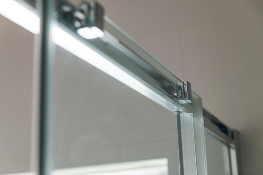 Glass shower door hinges. The top of the shower door. The door is made of glass and framed in metal. There is a chrome roller on top of the door.