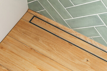 Shows a shower stall floor with a linear drain. The floor is made of wood-look tile and the drain is made of metal. The wall is tiled with light green subway tiles in a herringbone pattern.