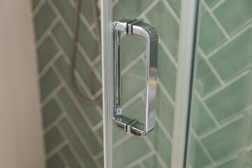 Silver Glass door handle of a glass partition shower unit. Bathroom glass door detail with bath tub in the background. Selective focus and soft light effect added.