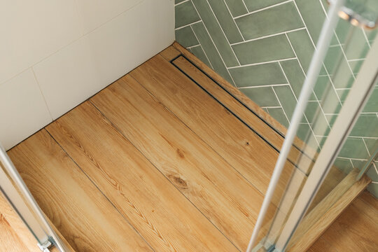 Shows a shower stall floor with a linear drain. The floor is made of wood-look tile and the drain is made of metal. The wall is tiled with light green subway tiles in a herringbone pattern.