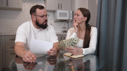 Young married couple sitting at glass kitchen table counting last remaining dollars, trying to figure out how to pay bills, woman thoughtfully holding finger on lips, man pointing at banknotes - Powered by Adobe