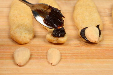 A close-up horizontal shot showing a spoonful of dark plum jam being applied to a baked witch finger cookie for attaching the almond nail
