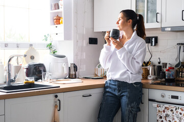 A woman drinks her morning coffee standing in the kitchen and looks out the window