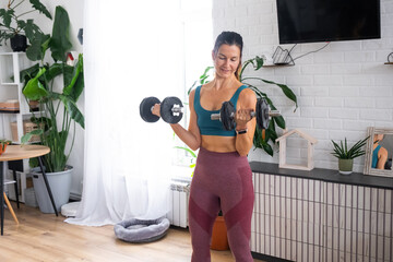 A woman in a sports uniform is engaged in fitness and strength training with a barbell and dumbbells at home in the interior