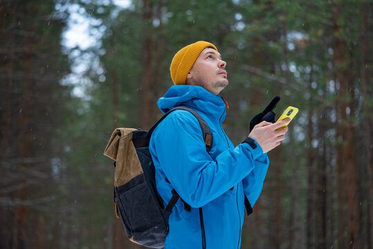 Young man wearing winter clothing and a backpack getting lost in a snow-covered forest, using a smartphone for navigation and looking up for orientation in deep nature