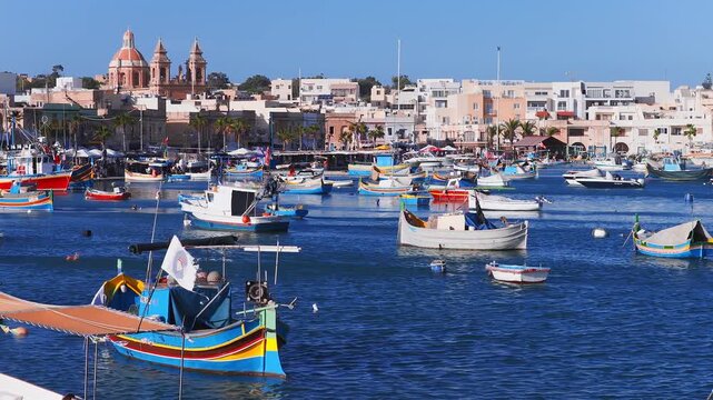 Aerial view of Marsaxlokk harbor in Malta shows luzzu and fishing boats on deep blue water, the seafront, and the terracotta domed parish church under midday light.