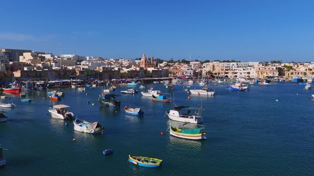 Aerial view of Marsaxlokk, Malta shows luzzu boats, twin domed church, and a busy promenade. Midday light, calm water, and people move along the quay.