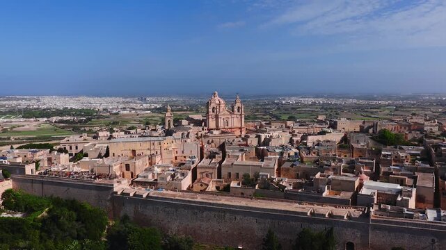 Aerial drone pans across Mdina, Malta, showing city walls, St. Pauls Cathedral, limestone rooftops, narrow streets, ramparts, and bastions in vivid daylight.