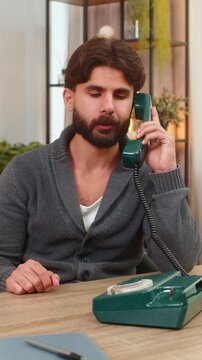 Indian man at home uses retro wired telephone smiles after short talk places handset back on base. Arabian guy at desk ends call friendly tone writes quick notes and returns to laptop for work tasks