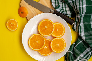 A top-down view shows orange slices on a white plate, a patterned knife on a cutting board, a whole orange, and a green checkered cloth, all on a bright yellow background.