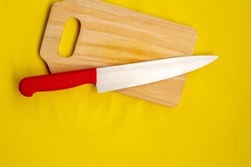 A top-down view of a kitchen knife with a red handle resting on a wooden cutting board, both set against a solid bright yellow background with copy space.