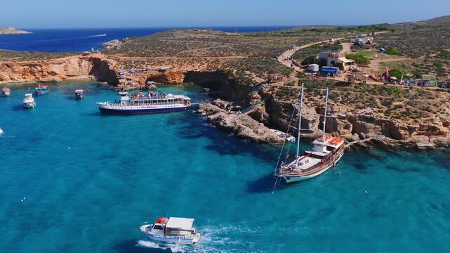 Aerial wide view shows Blue Lagoon at Comino, Malta. Hornblower tour boat, a wooden gulet, and small craft gather as tourists swim, sunbathe, and board in vivid light.