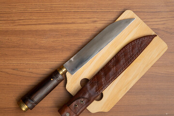 large, fixed-blade slaughter knife with a dark wooden handle and brass accents rests diagonally across a light wooden cutting board, next to its textured brown leather sheath.