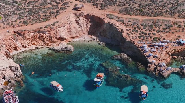 Aerial view of a sheltered cove on Comino, Malta, with reddish cliffs, umbrellas, boats, swimmers, snorkelers, and kayaks in clear blue green water at midday.