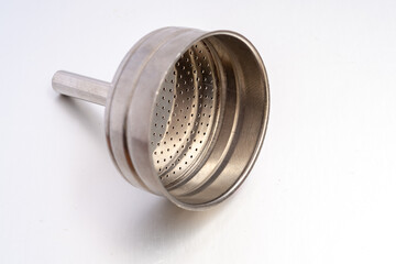A high-angle view of a used, stained aluminum filter funnel from a moka pot. It rests on its side on a white surface, showing filter holes and coffee ground residue.