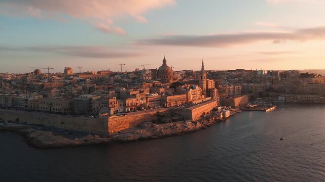 Aerial view of Valletta old town and Grand Harbour at sunset, with basilica dome, cathedral spire, boats and cars in warm light, wide framing and soft backlight.