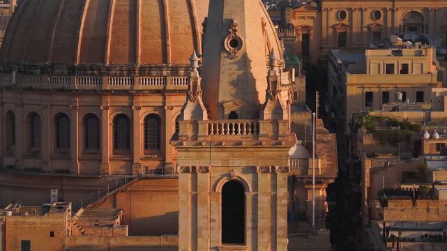 Aerial drone pans across a Baroque limestone dome and towers in Valletta, Malta at sunset, showing ribs, clerestory windows, balustrades, finials, and rooftop tanks.