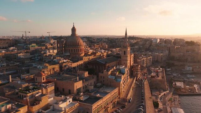 Aerial drone sweep tracks Valletta old town and Grand Harbour, domes and spires rise above limestone roofs and Maltese balconies as cars move and boats rest at sunset.