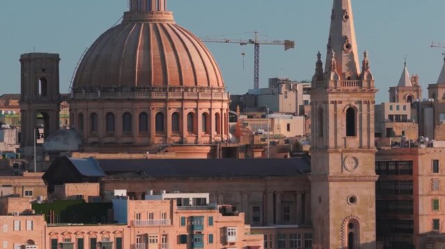 Aerial view shows Valletta, Malta at sunset, with the dome of Our Lady of Mount Carmel and the spire of St Paul's Pro Cathedral, rows of painted balconies, and a distant crane.