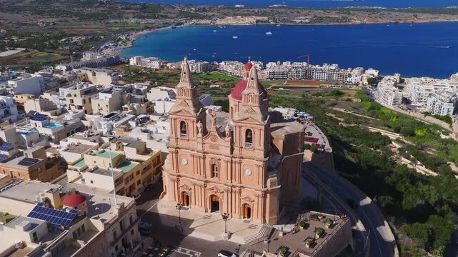 Aerial view arcs and descends over Mellieha parish church in Malta, twin towers and red dome, plaza and white buildings, boats and cars move under bright midday light.