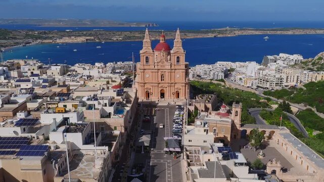 Aerial drone pass shows Mellieha parish church, red dome and towers, paved street to blue bay, boats crossing, Comino and Gozo on horizon, midday light and wide framing.