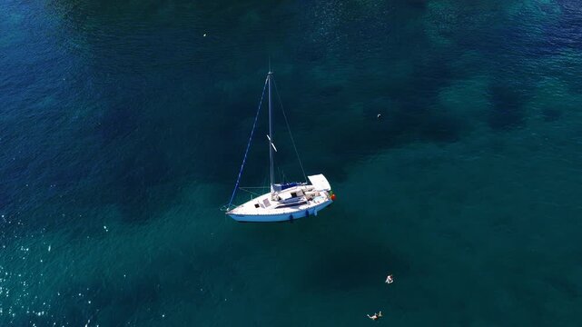 Aerial top down view shows a white sloop at anchor off Sliema, Malta, marked by an orange buoy, two swimmers drifting nearby, and glittering midday highlights.