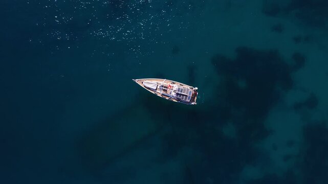 Aerial overhead view shows a wooden sailboat drifting off Sliema, Malta. A person in red is on deck. Bright sun casts mast shadow on shallow seabed below.