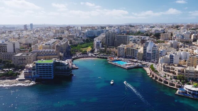 Aerial view of Balluta Bay in Sliema, Malta with boats, swimmers, a floating lido, and Balluta Parish Church at left in bright midday light and wide composition.
