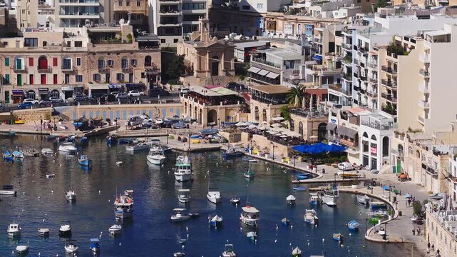 Aerial view of Sliema seafront in Malta shows a curved marina with luzzu boats, limestone townhouses, balconies, cafes, and people strolling in bright midday light.