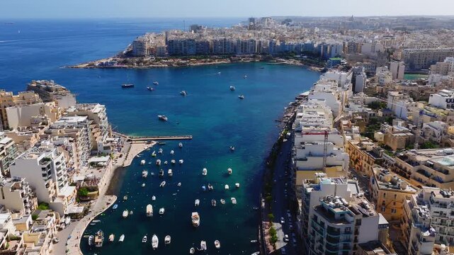 Aerial view of Balluta Bay in Sliema, Malta shows boats, a short pier, the Balluta Parish Church, limestone apartments, and Tower Road under bright midday light.