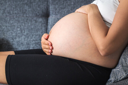 Close-up of pregnant woman gently holding her bare belly while sitting on sofa, showing love, care and anticipation of motherhood, concept of pregnancy, maternal health emotional connection with baby