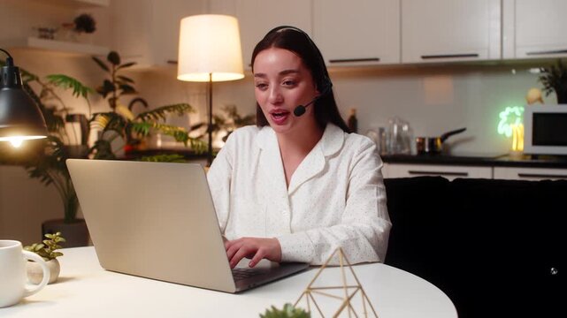 Young woman at night home works as call center agent with headset answers support call and logs ticket. Caucasian girl on sofa at evening listens speak note request polite tone service aid quick log.