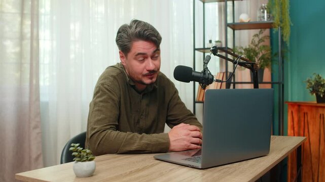 Middle-aged man at home records vlog speaking lively gestures toward laptop microphone engaging viewers online. Young adult guy at table shares ideas delivering content with passion through live show