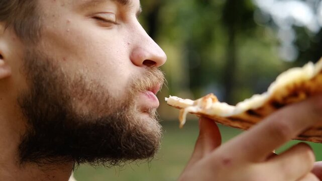 Bearded man enjoys eating a slice of pizza in the park on a sunny day. Relaxed outdoor lunch and casual lifestyle moment. 4k horizontal footage