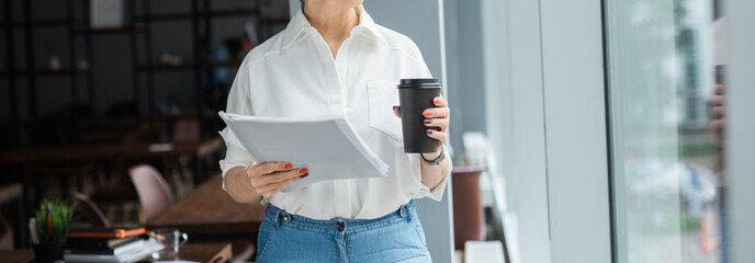 Woman standing by window with folder and coffee cup in hands. Banner with copy space. Professionalism, focus, and active work routine in modern lifestyle.