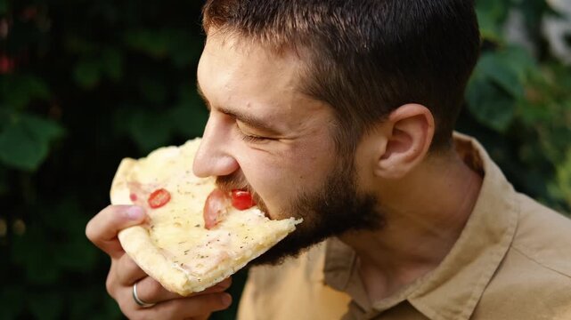 Close-up of a bearded man enjoying a slice of pizza with cheese, ham, tomato, and olives in the park on a sunny day. Relaxed summer vibe. 4k horizontal footage